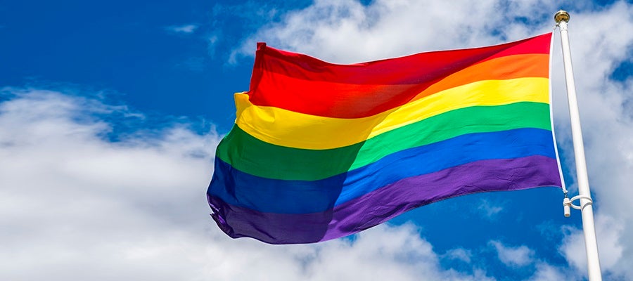 Gay Pride flag flying against a blue sky with white clouds.